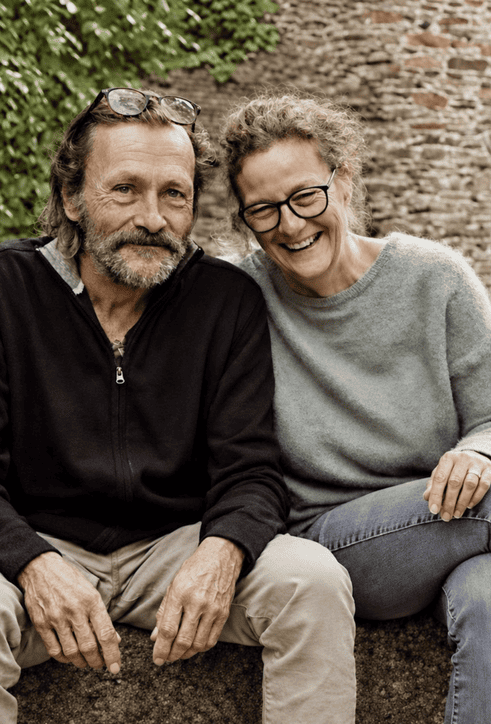 Older couple sitting close together on a stone ledge outdoors, smiling at the camera with a brick wall behind them.