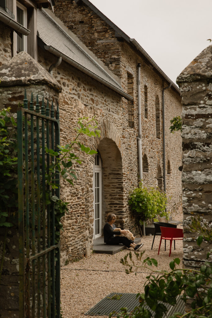 Older stone building with an arched entryway; a person sits on the steps with a dog in a gravel courtyard beside green and red chairs.