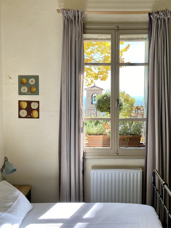 Cozy bedroom with white bedding and sunlight streaming through a large window, view of potted plants and the sea outside, beige walls and striped curtains.