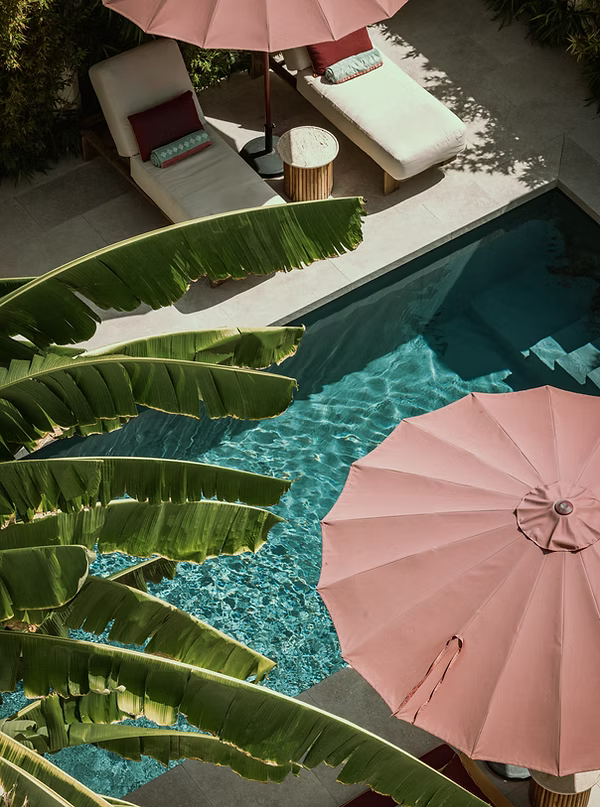 Overhead view of a turquoise pool surrounded by pink umbrellas, beige lounge chairs with cushions, and large banana leaves casting shade on the deck.